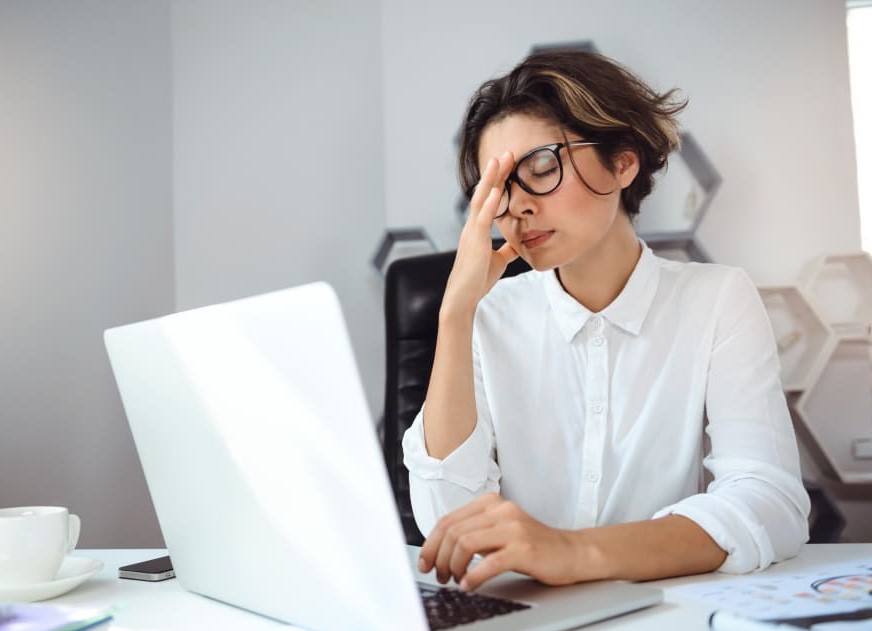 femme au travail, assise devant son ordinateur portant sa main à son visage qui semble fatiguée, surmenée